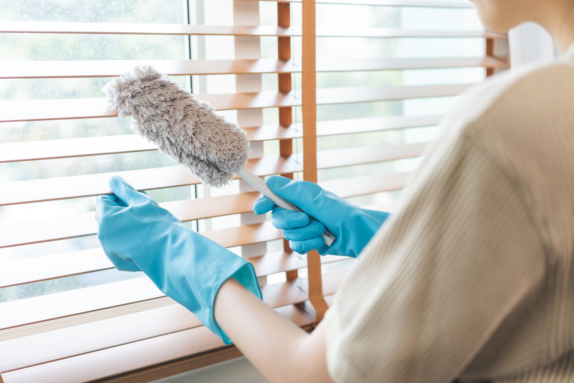 Eine Person mit blauen Handschuhen reinigt Jalousien mit einem Staubwedel.
